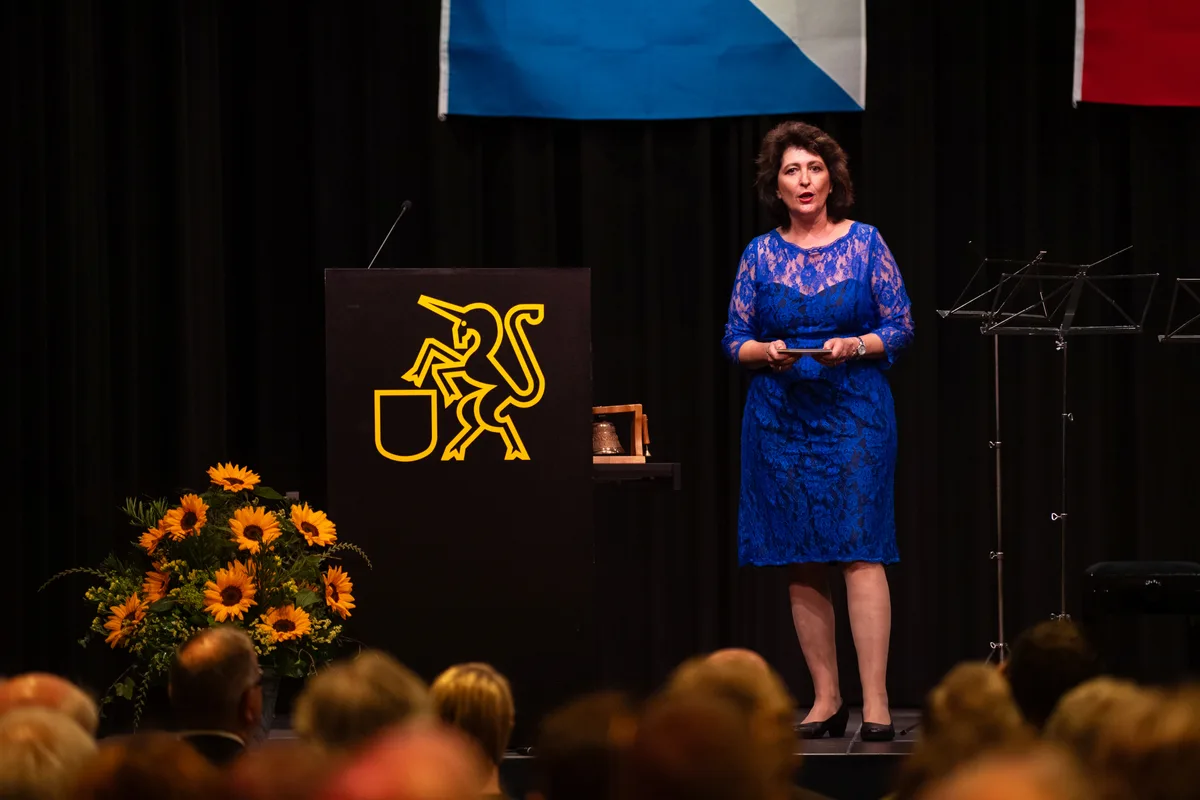 Eine Frau im einem blauen Kleid steht auf einer Bühne und spricht. 